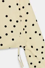 Chaqueta de cuello subido y manga larga con estampado de lunares negros sobre base beige. Cuenta con bolsillos de vivo en los laterales, detalles de frunces ajustables con cordones en el delantero y cierre frontal con botones.