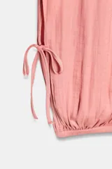 Pantalón de mujer color beige claro, de tiro medio y calce holgado, confeccionado en tejido ligero y fluido. Presenta cintura elástica ajustable con lazada, bolsillos laterales y aberturas ajustables con lazadas en las piernas que terminan en bajo abullonado con elástico.