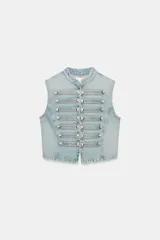 Chaleco de denim color celeste con cuello subido, sin mangas, con detalle de aplicaciones horizontales y botones metálicos en el frente. Cuenta con cierre frontal con ganchos metálicos ocultos.