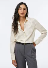Camisa de corte clásico y entallado, color beige con rayas verticales finas en color negro. Presenta cuello camisero, cierre frontal con botones y mangas largas con puños abotonados.