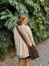 Bolso tipo bandolera de gran tamaño, confeccionado en gamuza color marrón. Presenta una correa ancha ajustable con hebilla metálica y costuras visibles en el cuerpo del bolso.