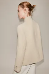 Blazer de corte estructurado en color beige, con cuello mao y cierre frontal cruzado mediante botones. Presenta mangas largas con detalle de botones en los puños y bolsillos de vivo en el frente.