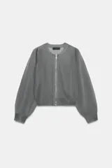 Chaqueta estilo bomber de punto color beige con acabado brillante, cuello redondo, manga larga con puños elásticos y cierre frontal con cremallera.