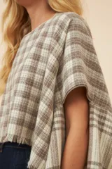 Poncho color beige con diseño a cuadros, cuello bote y terminaciones desflecadas. Tiene aberturas para los brazos en los laterales y detalles en cuero.