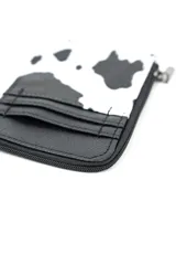 Billetera tarjetero con diseño estampado tipo animal print de vaca en blanco y negro, con sección inferior en cuerina negra que incluye ranuras para tarjetas. Posee cierre lateral, herrajes metálicos y una pequeña cadena con argolla para llavero.