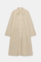 Trench largo de Zara Woman Collection, confeccionado en color beige. Cuenta con cuello de solapa, manga larga, bolsillos frontales de parche y cierre central con botones visibles.
