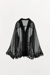 Blusa semitransparente negra de cuello solapa y manga larga, con cierre frontal de botones y detalles de bordados tonales en el ruedo y los puños.