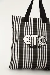 Bolsa tote de diseño amplio a rayas verticales en blanco y negro, con asas de hombro negras y aplique frontal bordado con letras.