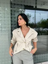 Chaqueta color nude con cuello solapa, cierre frontal de botones dorados y bolsillos laterales. Presenta detalles de pliegues horizontales en los hombros y verticales en la cintura.