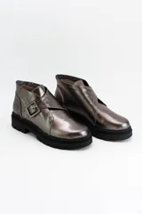 Botines de cuero metalizado en tono oscuro con acabado brillante. Presentan un diseño de corte bajo con una hebilla lateral decorativa y suela de goma negra con plataforma dentada.