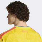 Camiseta de fútbol Adidas de la selección de Colombia, modelo Primera Equipación 26, color amarillo con un patrón sutil en relieve. Presenta cuello en V con ribete azul, logo de Adidas en azul y escudo de la federación colombiana. Las tres franjas de Adidas en los hombros son rojas y azules.