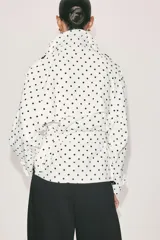 Cazadora estilo trench corta confeccionada en lino, con diseño de lunares negros sobre fondo blanco. Cuenta con cuello solapa, manga larga con trabillas en los puños, cierre frontal cruzado con botones negros y cinturón ajustable del mismo tejido.