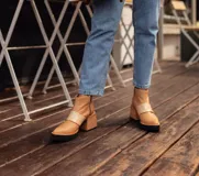 Botinetas de cuero color tostado con diseño de punta fina y taco cuadrado de madera. Presentan una banda horizontal de charol en tono más claro sobre el empeine y suela de goma negra.