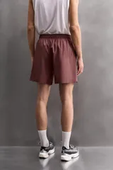 Pantalones cortos deportivos color marrón rojizo, confeccionados en tejido técnico ligero y elástico. Presentan cintura elástica ajustable con cordón interior, bolsillos frontales y un bolsillo trasero con cierre de cremallera. Los laterales tienen perforaciones para transpirabilidad y aberturas laterales en el bajo.