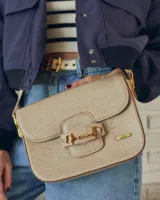 Cartera tipo bandolera de cuero con textura grabada, color beige. Presenta un detalle de herraje metálico dorado en la solapa frontal y placa con logo de la marca. Incluye correa ajustable para llevar al hombro o cruzada.