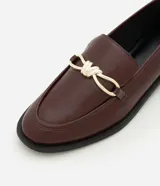 Mocasines de mujer confeccionados en poliuretano color marrón oscuro, con acabado brillante y detalle decorativo metálico dorado en el empeine. Presentan una silueta clásica de punta redondeada y suela plana con pequeño tacón.