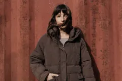Campera larga acolchonada color marrón, con diseño midi, capucha amplia, cierre frontal con cremallera y broches, y bolsillos delanteros con tapa.