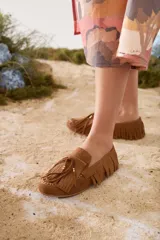 Mocasines de gamuza color tostado, con diseño clásico, flecos alrededor del calzado y en la capellada, y cordón decorativo con detalles metálicos dorados.