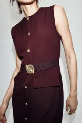 Vestido midi de cuello redondo y sin mangas, confeccionado en tejido tipo crepe. Presenta cierre frontal con botones dorados, bolsillos con solapa en la parte delantera y un cinturón elástico integrado con hebilla metálica decorativa.