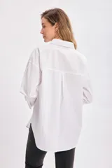 Camisa blanca de manga larga con cuello clásico y botones frontales. Presenta un corte holgado (oversize) y un bolsillo de parche en el pecho izquierdo.