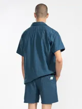 Bermuda de hombre color azul petróleo, confeccionada en mezcla de lino y algodón. Presenta cintura elástica con cordón, bolsillos laterales y trasero tipo parche. Incluye bordado floral en la pierna izquierda y etiqueta con logo en el bolsillo trasero.