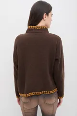 Sweater de punto color marrón con cuello alto, manga larga y calce relajado. Presenta detalles bordados en color mostaza en el cuello, puños, ruedo y bolsillo frontal, además de un aplique en forma de corazón en el bolsillo.