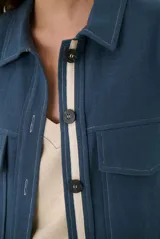 Sobrecamisa de lino en color azul, con diseño de corte recto, cuello camisero, cierre frontal con botones oscuros y detalle de cinta en contraste a lo largo de la botonadura. Cuenta con bolsillos frontales con solapa y costuras visibles en color claro.