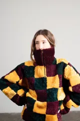 Buzo de cuello alto con cierre frontal parcial, confeccionado en tejido de pelo sintético con diseño de cuadros grandes en colores amarillo, bordó y verde oscuro. Presenta un corte holgado y bolsillos laterales con detalles en cuero.