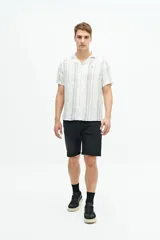 Camisa de manga corta con cuello cubano, confeccionada en una mezcla de lino y algodón con textura. Presenta un diseño a rayas verticales en tonos beige y celeste sobre fondo blanco, con cierre frontal de botones y logo bordado en el pecho.