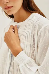 Camisa de algodón blanco con textura de pequeños puntos en relieve, cuello redondo con cierre de botones oculto, detalles de alforzas verticales y encaje en el frente, y mangas largas con puños elásticos.