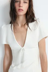 Vestido corto de Zara, color blanco, con escote en pico, mangas cortas abullonadas y detalle de lazo frontal en el mismo tejido. Presenta un corte entallado y cierre de cremallera oculta en la espalda.