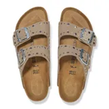 Sandalias Birkenstock modelo Arizona con dos tiras ajustables de gamuza color taupe, decoradas con remaches metálicos plateados. Presenta plantilla anatómica de corcho y suela de goma negra.