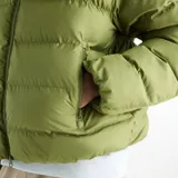 Campera puffer New Balance modelo Boston, color verde oliva, con capucha, cierre frontal completo y logo blanco estampado en el pecho.