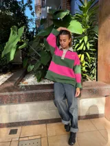 Buzo de tejido de punto con diseño de rayas horizontales anchas en color verde y fucsia. Presenta cuello tipo polo en color crema con cierre frontal de botones, mangas largas y aberturas laterales en el ruedo.