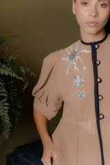 Chaqueta gris de silueta femenina con mangas abullonadas, cuello redondo y cierre frontal con botones forrados. Presenta detalles de bordado artesanal a mano en los hombros y solapas.