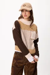Sweater de punto con diseño patchwork en tonos gris, grafito y crudo, con cuello alto tejido. Presenta una estructura de bloques de color y texturas variadas, incluyendo detalles de trenzas y tejido acanalado.