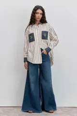 Camisa de corte oversize con estampado de rayas verticales, cuello clásico y cierre frontal de botones. Presenta dos bolsillos frontales en contraste con bordados de tréboles, además de puños y cuello en color sólido con detalles en amarillo.