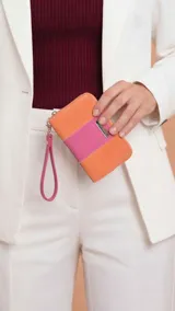 Billetera mediana de cuero sintético con diseño bicolor en naranja y fucsia. Cuenta con cierre perimetral, correa de muñeca extraíble y placa metálica decorativa con logo.