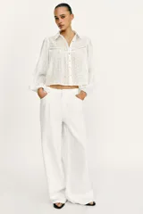 Blusa corta de color blanco con diseño de bordados perforados, cuello solapa, cierre frontal con botones y mangas largas abullonadas.