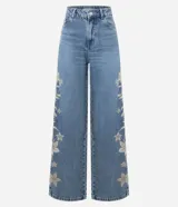 Pantalón wide leg de denim azul con bordados florales en los laterales. Presenta cintura baja, cierre con botón y cremallera, y bolsillos funcionales.