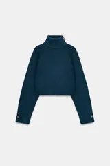 Jersey de punto de tacto suave, color azul petróleo, con cuello subido y manga larga. Presenta detalle de botones metálicos en el hombro y en los puños.