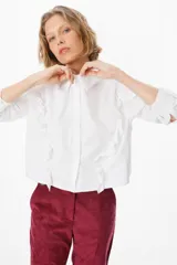 Camisa de corte cropped en color azul, con cuello clásico y cierre frontal de botones. Presenta volados decorativos que recorren la parte frontal y las mangas largas, terminando en puños abotonados.