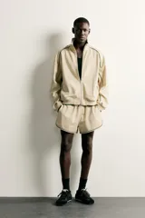 Chaqueta estilo deportivo de corte relajado, color beige con detalles de vivos en color verde oscuro. Presenta cuello alto, cierre frontal con cremallera de doble dirección, bolsillos laterales y puños elásticos.