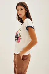 Remera de algodón color crema con cuello y bordes de mangas en contraste azul marino. Presenta un diseño frontal con bordados de flores coloridas y la inscripción 'MARCHÉ aux FLEURS' en letras azules.