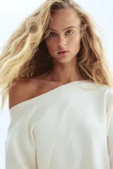 Buzo corto de punto fino en color crema, con diseño de un solo hombro y escote asimétrico. Presenta mangas largas y puños acanalados.