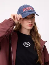 Gorra con visera curva de estilo clásico, confeccionada en algodón con efecto lavado. Presenta el logo de la marca Rusty bordado en el frente con letras en color rojo y borde blanco.