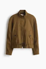 Campera corta verde militar en mezcla de lino, con cuello elevado, mangas raglán, bolsillos frontales con solapa y botones, y cordón ajustable en el bajo.