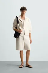 Bermuda de corte relajado color beige claro, confeccionada en tejido con textura. Presenta cierre frontal con cremallera y botón, bolsillos delanteros y bolsillos traseros de doble vivo con botón.