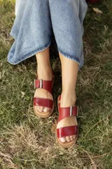 Sandalias de cuero rojo con dos tiras amplias en el empeine, una lisa y otra con hebilla metálica rectangular, y suela plana.