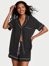 Pijama corto negro con estampado de lunares blancos, compuesto por una camisa de manga corta abotonada con ribetes blancos y un short a juego con cordón de ajuste rosado.